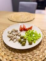 Plate of fresh ingredients featuring chopped chicken, olives, sliced green bell peppers, and quartered tomatoes on a wicker placemat. Healthy and appetizing meal preparation setting photo