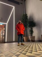 Millennial man stands confidently in a modern space, wearing a vibrant orange parka, surrounded by geometric patterns and ambient lighting. Blend of contemporary style and cozy warmth photo