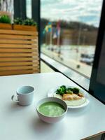 Healthy meal featuring a bowl of green soup and a plate with broccoli and fish is set on a white table by a large window. Peaceful dining atmosphere photo