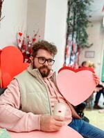 Smiling man holds a large pink heart on Valentine day while sitting at festively decorated table, capturing spirit of love and warmth in a cozy setting. Cheerful festive atmosphere evoke photo