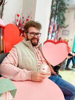 Smiling man holds a large pink heart on Valentine day while sitting at festively decorated table, capturing spirit of love and warmth in a cozy setting. Cheerful festive atmosphere photo