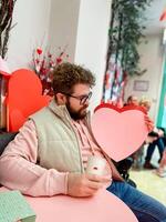 Smiling man holds a large pink heart on Valentine day while sitting at festively decorated table, capturing spirit of love and warmth in a cozy setting. Cheerful festive atmosphere evoke feelings photo