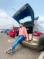 Millennial man in a pink hoodie sits casually on the edge of an open car trunk in a parking lot. Relaxed and contemplative, enjoying a moment of pause under the wide sky photo