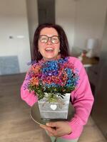 Cheerful millennial woman with glasses is joyfully holding a colorful bouquet of flowers in a decorative pot. Pink sweater and radiant smile add warmth and vibrancy to the scene photo