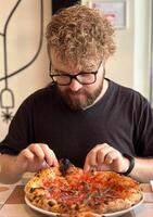 Millennial man with curly hair and glasses looking on a delicious-looking pizza in restaurant. Moment of culinary anticipation and simple joys of food photo