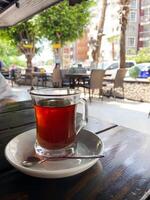Glass of tea on a white saucer with a spoon, set on an outdoor cafe table with a view of a bustling urban street scene in the background. Essence of a relaxing tea moment amidst city life photo