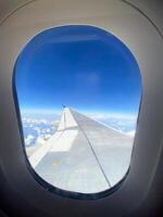 View of airplane wing through window at high altitude, showcasing clear blue sky and clouds far below. Serene beauty of flight and the expansive view of the horizon from above. photo