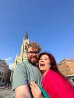 Funny couple taking a selfie in front of a historical building on a sunny day, with clear blue sky and impressive architecture. capturing moments of travel and togetherness photo