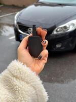 Woman's hand holding a car key fob in front of a black modern car on a rainy day. Fluffy jacket highlights the close-up shot of the key, ready to unlock the vehicle. photo