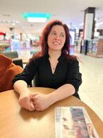 Cheerful millennial woman sits at a table in a bustling shopping mall, exuding warmth and friendliness. Joyful atmosphere of shared moments and relaxation photo