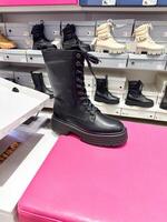 Black leather combat boot displayed on a bright pink bench in a shoe store. Various other boots in different colors and styles are neatly stacked in the background photo