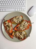 Delicious stuffed peppers with melted cheese and herbs on a plate, placed near a computer keyboard and mouse on a white desk. Perfect blend of work and healthy eating photo