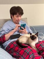 Elderly woman using smartphone and social networks with a siamese cat on their lap, relaxing on a couch in cozy indoor photo