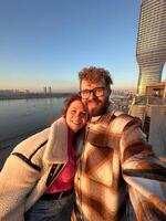 Couple taking selfie on a balcony, overlooking a river with a modern skyscraper in Belgrade city during a serene sunset. the warm glow of the evening light casts photo