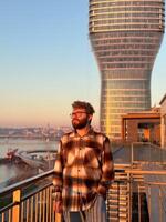 Man stands thoughtfully on a balcony, overlooking a river with a modern skyscraper in Belgrade city during a serene sunset. the warm glow of the evening light casts photo
