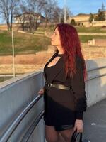 Millennial woman stands confidently in sunlight, with vibrant red hair flowing and dressed in a chic black outfit. Background of a clear blue sky and distant scenery enhances the dynamic photo