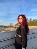 Millennial woman stands confidently in sunlight, with vibrant red hair flowing and dressed in a chic black outfit. Background of a clear blue sky and distant scenery enhances the dynamic photo