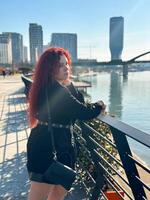 Millennial woman with long curly red hair leans on a black railing, gazing thoughtfully at the camera on a sunny day. Background features modern buildings and a river, evoking a sense of urban calm photo