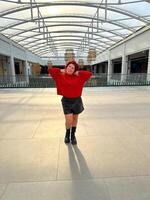Millennial woman poses confidently in an open, modern environment, wearing a vibrant red sweater and black boots. photo