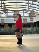 Millennial woman poses confidently in an open, modern environment, wearing a vibrant red sweater and black boots. photo