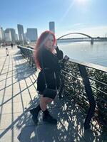 Millennial woman with long curly red hair leans on a black railing, gazing thoughtfully at the camera on a sunny day. Background features modern buildings and a river, evoking a sense of urban calm photo
