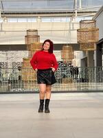 Millennial woman poses confidently in an open, modern environment, wearing a vibrant red sweater and black boots. photo