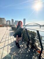 Millennial woman with long curly red hair leans on a black railing, gazing thoughtfully at the camera on a sunny day. Background features modern buildings and a river, evoking a sense of urban calm photo