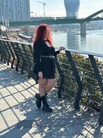 Millennial woman with long curly red hair leans on a black railing, gazing thoughtfully at the camera on a sunny day. Background features modern buildings and a river, evoking a sense of urban calm photo