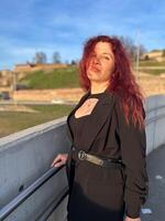 Millennial woman stands confidently in sunlight, with vibrant red hair flowing and dressed in a chic black outfit. Background of a clear blue sky and distant scenery enhances the dynamic photo