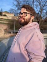 Millennial man enjoys a peaceful moment autumn outdoors, wearing a soft pink hoodie and glasses. Background of trees and historic architecture creates an atmosphere of tranquility and contemplation photo