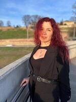 Millennial woman stands confidently in sunlight, with vibrant red hair flowing and dressed in a chic black outfit. Background of a clear blue sky and distant scenery enhances the dynamic photo