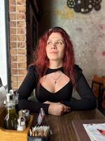 Millennial woman with red hair sits thoughtfully at a rustic cafe table, surrounded by a cozy and artistic atmosphere. Soft lighting and her pensive expression convey photo