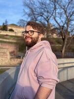 Millennial man enjoys a peaceful moment autumn outdoors, wearing a soft pink hoodie and glasses. Background of trees and historic architecture creates an atmosphere of tranquility and contemplation photo