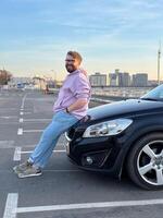 Millennial man in a casual hoodie leans confidently against a sleek black car in a parking lot. Peaceful evening sky forms a serene backdrop to this moment of relaxation and contemplation photo