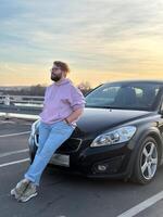 Millennial man in a casual hoodie leans confidently against a sleek black car in a parking lot. Peaceful evening sky forms a serene backdrop to this moment of relaxation and contemplation photo
