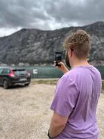 Millennial man standing by a car and take photo on smartphone, capturing the scenic view of a mountain range with his camera phone for social networks blog.