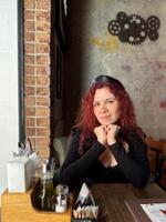 Millennial woman with red hair sits thoughtfully at a rustic cafe table, surrounded by a cozy and artistic atmosphere. Soft lighting and her pensive expression convey photo