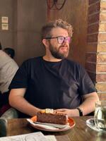 Bearded curly millennial man sitting in a cozy cafe with a slice of cake in front, enjoying a quiet moment in a rustic setting. photo