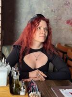 Millennial woman with red hair sits thoughtfully at a rustic cafe table, surrounded by a cozy and artistic atmosphere. Soft lighting and her pensive expression convey photo