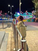 Millennial woman in a fluffy jacket stands illuminated by the colorful lights of a carnival ride in amusement park, gazing upwards with a joyful expression. Winter festive atmosphere photo