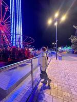 Millennial woman in a fluffy jacket stands illuminated by the colorful lights of a carnival ride in amusement park, gazing upwards with a joyful expression. Winter festive atmosphere photo