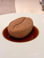 Close-up delicately crafted dessert resembling a coffee bean rests on a plate, surrounded by a rich brown sauce. Creative culinary presentation combines texture photo