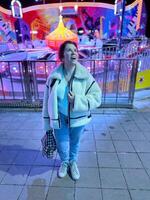 Millennial woman in a fluffy jacket stands illuminated by the colorful lights of a carnival ride in amusement park, gazing upwards with a joyful expression. Winter festive atmosphere photo