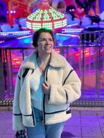Millennial woman in a fluffy jacket stands illuminated by the colorful lights of a carnival ride in amusement park, gazing upwards with a joyful expression. Winter festive atmosphere photo
