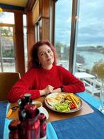 Millennial woman in a red sweater sits at a restaurant table by the window. In window scenic river view enhances the calm and reflective mood. Breakfast in hotel cafe photo