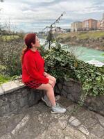 Millennial woman in a red sweater sitting on a stone ledge, enjoying a tranquil river view with a backdrop of city buildings. Autumn scene photo