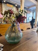 bouquet of colorful flowers is elegantly arranged in a glass vase on a rustic wooden table in a cozy cafe. the backdrop of the cafe adds warmth and charm to the serene scene photo