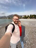 couple takes a cheerful selfie on a pebbled beach, with vast mountains and a serene sea in the background. their relaxed expressions and casual attire complement the picturesque coastal scenery photo