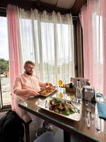man in a relaxed setting enjoys a meal at a glass table, surrounded by soft pink curtains with a view outside. a delicious dish is elegantly presented in front of him photo