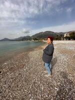Millennial woman stands alone on a pebbled beach, gazing out at the tranquil sea under a partly cloudy sky. the serene setting is framed by distant mountains and a coastal cityscape photo